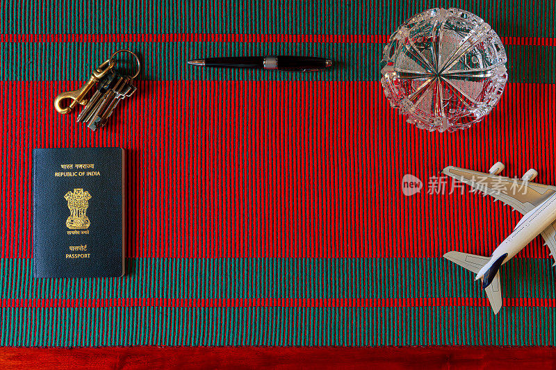 平坦的Lay For Travel - The New India Passport And A Group Of Objects Placed On A Woven Fabric Of Red And Green Hinting At Christmas - A圆珠笔，钥匙和一架模型飞机。一个水晶烟灰缸为图像增添了一些活力。